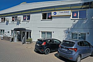 Ein weißes Gebäude mit dem Schild Trans Global Logistics Europe GmbH über dem Eingang. Zwei Autos sind auf dem Parkplatz davor geparkt.