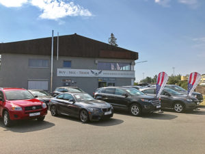 Autohaus mit mehreren ausgestellten Autos auf einem Parkplatz, darunter ein rotes, ein schwarzes und ein graues Fahrzeug. Das Gebäude im Hintergrund hat ein Schild mit „Buy - Sell - Trade“. Im Vordergrund stehen Flaggen. Der Himmel ist leicht bewölkt.
