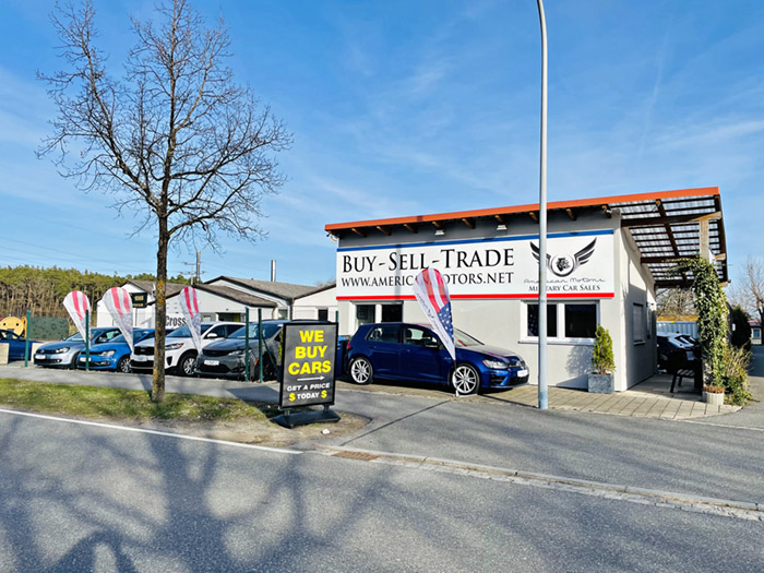 Autohandelsgeschäft mit Werbeschildern Buy - Sell - Trade und We Buy Cars vor dem Gebäude. Vor dem Geschäft stehen mehrere Autos und bunte Fahnen. Ein Baum und eine Straße sind im Vordergrund zu sehen.