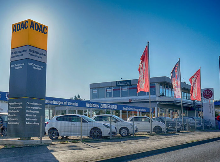 ADAC-Zentrum und Autohaus Zimmermann mit mehreren weißen Autos im Vordergrund. Mehrere rote Flaggen wehen vor dem Gebäude, und ein großes gelbes ADAC-Schild steht links im Bild.