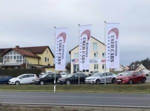 Gebrauchtwagenplatz mit mehreren Autos, drei Fahnen mit der Aufschrift Used Cars und Häusern im Hintergrund.