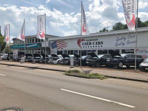 Gebrauchtwagenhändler mit dem Namen Ramstein Used Cars. Mehrere Autos sind vor dem Gebäude ausgestellt, mit amerikanischen Flaggenmotiven auf den Bannern. Es gibt Schilder mit der Aufschrift Quality Cars, Quality Service.