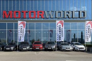 Mehrere Autos stehen vor dem Gebäude von Motorworld, das große Glasfenster und Werbefahnen zeigt.