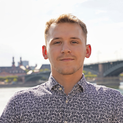 Cedric Hupfauf: Ein junger Mann mit kurzem Haar und gemustertem Hemd steht draußen vor einer verschwommenen städtischen Kulisse mit Fluss und Brücke im Hintergrund.