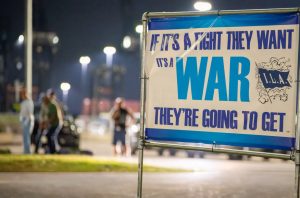 Ein Schild bei Nacht zeigt: If it's a fight they want, it's a war they're going to get. Im Hintergrund sind verschwommen Menschen und Lichter zu sehen.