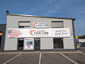 Grau gestrichenes Gebäude mit zwei großen Werbeschildern für Ramstein Wash Park und Ramstein Used Cars. Ein Schild zeigt die US-Flagge und der Text lautet Quality Cars, Quality Service. Vor dem Gebäude befinden sich einige Parkplätze.