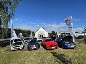 Eine Reihe von Autos steht auf einem Rasen vor einem Autohaus. Einige Fahrzeuge haben ihre Motorhauben geöffnet. Flaggen mit dem Logo des Unternehmens flankieren die Autos. Der Himmel ist klar und blau.
