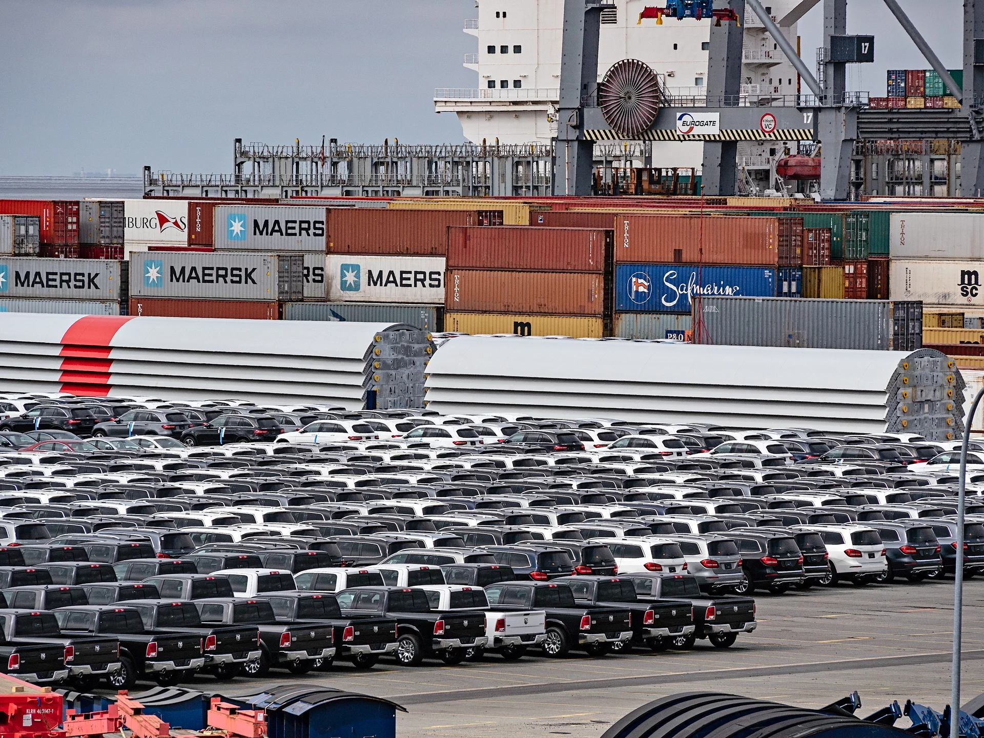 Reihen von geparkten Autos auf einem Hafengelände, im Hintergrund sind gestapelte bunte Container und große Hafenkräne zu sehen.