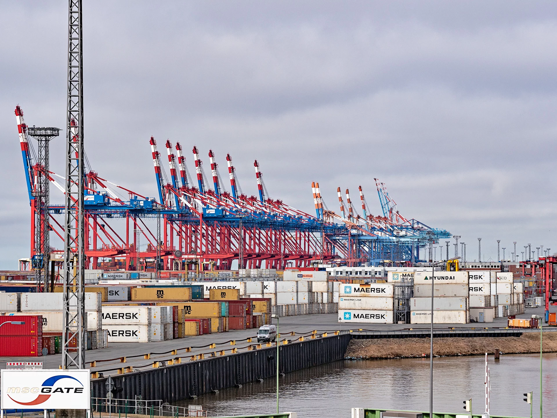 Hafenansicht mit zahlreichen Containern und rot-blauen Kränen entlang des Kais, bei bewölktem Himmel.