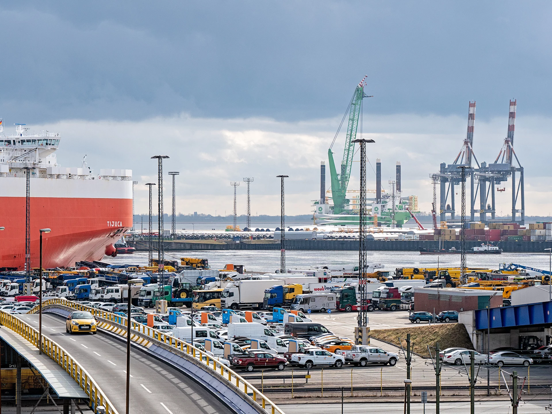 Ein Hafen mit einem großen Frachtschiff im Vordergrund, vielen geparkten Autos und LKWs, sowie Kränen und Containern im Hintergrund unter bewölktem Himmel.