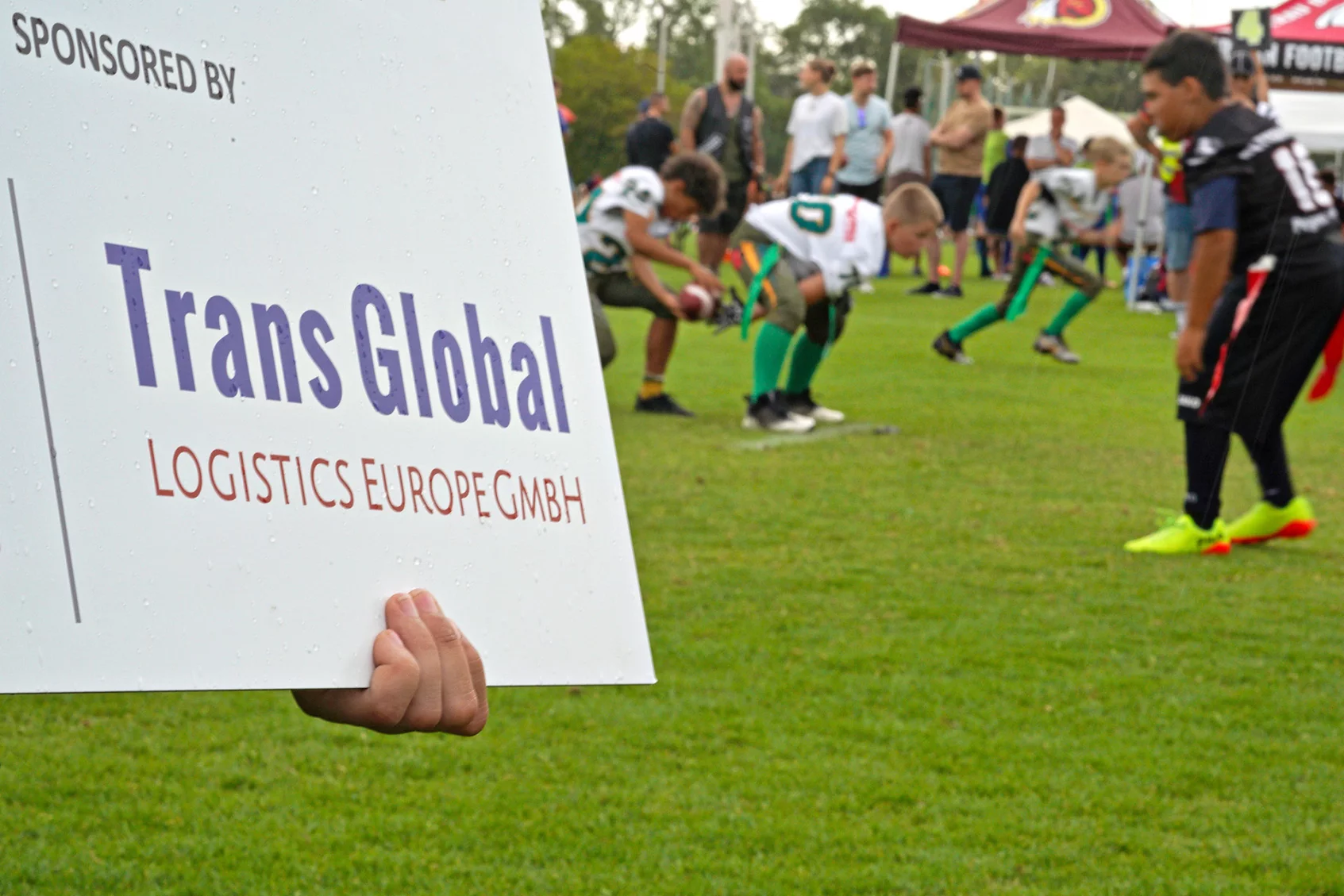 Schild mit der Aufschrift Sponsored by Trans Global Logistics Europe GmbH im Vordergrund; im Hintergrund Kinder, die sich auf ein Sportereignis vorbereiten, vermutlich American Football.